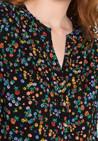 Blouse fleurie noire présentant un mélange de petites fleurs colorées et de feuilles vertes. Col en V avec trois boutons décoratifs à l'avant.