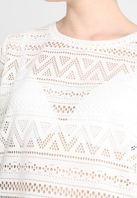 Bílá košile s propracovaným děrováním a geometrickými tvary. Měkká tkanina, kulatý výstřih a krátké rukávy.