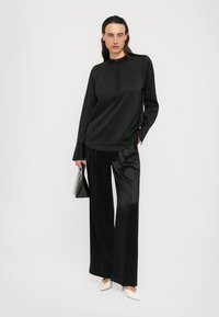 Person wearing black long-sleeve blouse and wide-leg satin pants, holding a black handbag, and white pointed heels against a plain white background.