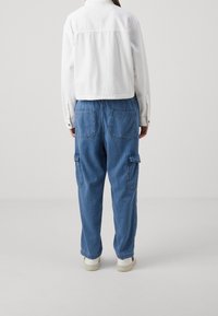 Vit, kort klädd denimjacka med knappstängning och detalj på baksidan, tillsammans med löst sittande blå cargobyxor med två bakfickor.