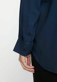 Chemise à manches longues bleu marine avec un poignet boutonné, dotée d'un petit détail brodé et d'une texture de tissu lisse.