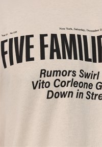 Camiseta de color beige claro con texto negro en negrita. Presenta un encabezado "CINCO FAMILIAS" y subtítulos. Material de algodón, diseño estándar de cuello redondo.