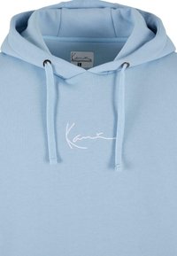 Svijetloplava dukserica s kapuljačom na vezanje, koja ima bijeli izvezeni logo na prsima. Izrađena od mekane tkanine s glatkom teksturom.