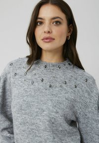 Pull gris avec col rond, orné de cristaux variés sur les épaules. Texture douce et coupe décontractée.
