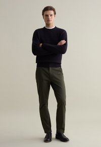 Marineblauwe, gestructureerde trui van breiwerk met geribbelde manchetten, gecombineerd met donkergroene, taps toelopende broeken en zwarte instapschoenen. Eenvoudig, minimalistisch ontwerp.