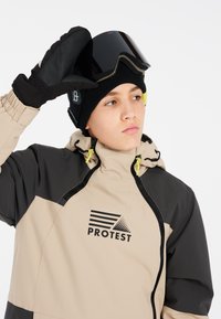 Giacca da neve beige e nera con cappuccio con zip, caratterizzata da un logo e dettagli giallo brillante. Guanti neri e un casco scuro completano il look.