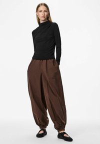 Czarna bluzka z długim rękawem i marszczonym dekoltem, zestawiona z luźnymi, brązowymi spodniami z elastycznym pasem. Całość uzupełniają czarne wsuwane buty.