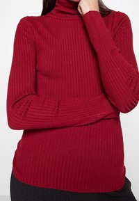 Pull à col roulé rouge côtelé à manches longues. Présente une coupe ajustée et un tissu texturé, s'étendant jusqu'à un ourlet subtil à la taille.