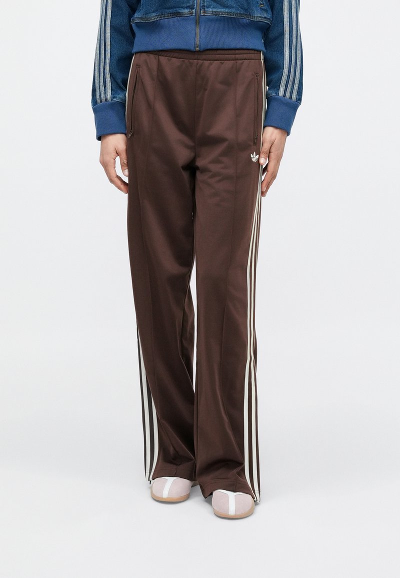 Pantalones deportivos marrones hechos de tela suave, con rayas blancas en los costados y dos bolsillos con cremallera. Combinados con zapatos grises sin cordones.