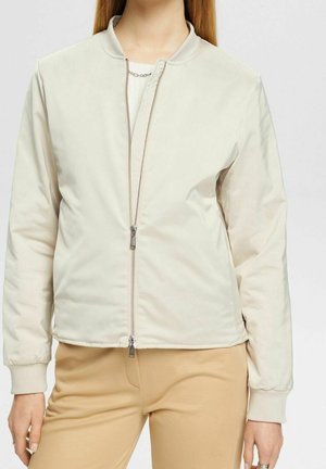 Femme portant une veste bomber beige clair à fermeture éclair avant, manches côtelées, et un pantalon beige, debout devant un fond uni.