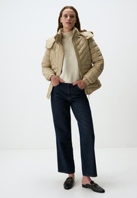 Beige gewatteerde pufferjack met een hoge kraag, gecombineerd met een lichtbeige sweater en donkerblauwe wijde jeans, afgewerkt met zwarte instappers.