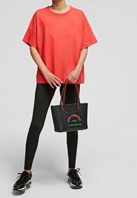 Korall oversized t-shirt parret med sorte leggings. Den sorte tote bag har orange håndtag og grøn accenttråd med logodetaljer.