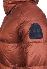 Giacca imbottita color arancio bruciato, con design trapuntato e una toppa blu scuro con logo sulla spalla. Texture liscia e lucida.