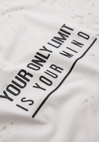 Tela de algodón blanca con un gráfico de texto negro que dice "TU ÚNICO LÍMITE ERES TÚ MISMO", con patrones de salpicaduras visibles en la superficie.