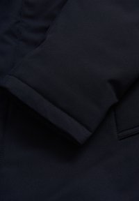 Navy sininen ulkovaatetuskangas, jossa on sileä, matta rakenne. Ominaisuuksiin kuuluu taitettu hiha ja näkyvät ompeleet kestävyyden takaamiseksi. Ei kuvioita tai koristeita.