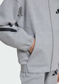 Sudadera con cremallera en color gris con acentos en negro y bolsillos laterales. La tela es suave con un acabado ligeramente texturizado. Los puños son elásticos.
