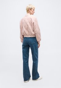 Άτομο με κοντά ξανθά μαλλιά που φοράει ανοιχτό ροζ επενδεδυμένο μπουφάν, μπλε τζιν με ίσια μπατζάκια και λευκά αθλητικά παπούτσια, στέκεται μπροστά σε λευκό φόντο.