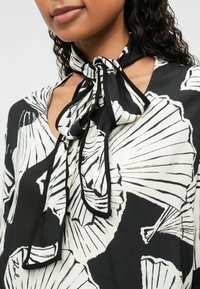 Blusa bianca e nera con motivo floreale a conchiglia, caratterizzata da un dettaglio a sciarpa annodata sul collo e un tessuto leggero e testurizzato.