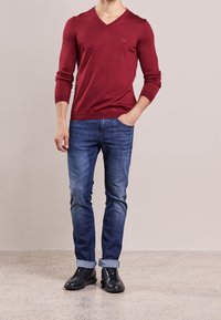 Röd V-ringad tröja i finstickad kvalitet, kombinerad med urtvättade blå jeans och svarta sneakers. Tröjan har en diskret logotyp på bröstet och en figurnära passform.