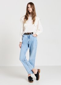 Blouse blanche à volants avec des détails de rayures verticales, jeans droite bleu clair, et sabots en bois noirs, avec une semelle beige et des clous dorés.