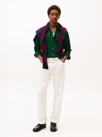 Grüne Knopfhemd, weiße Hose, schwarze Schuhe und ein kariertes navy- und burgunderfarbenes Sweatshirt über den Schultern. Einfarbiger Hintergrund.