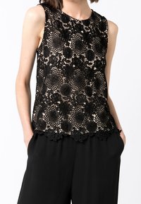 Svart spets ärmlös topp med blomsterdesign, intrikata mönster, rundad halsringning och vågig nederkant, lagd över en beige bakgrund.