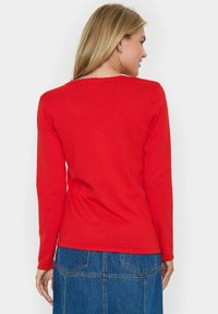 Pull rouge à manches longues avec un col rond, texture côtelée et ourlet à volants. Porté avec une jupe en denim et vu de dos.