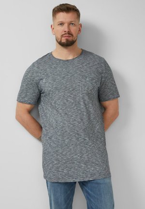 Man met baard, gekleed in een grijs, kort mouw T-shirt met gemêleerde stof en een blauwe spijkerbroek, staand tegen een lichtgrijze muur met zijn handen achter zijn rug.