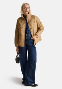 Gepatchte beige Jacke mit hohem Kragen, kombiniert mit einer dunkelb blauen weit geschnittenen Jeans. Eine schwarze Handtasche betont das Outfit und schafft einen schichtartigen Look.