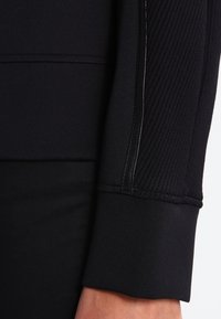 Gros plan d'une manche longue noire en tissu texturé avec un poignet lisse sur le bras d'une personne sur un fond clair uni.