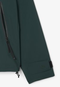 Donkergroene jack met een zwarte ritssluiting, verstelbare manchetten met klittenbandsluiting en een subtiele geborduurde logo nabij de manchetten.