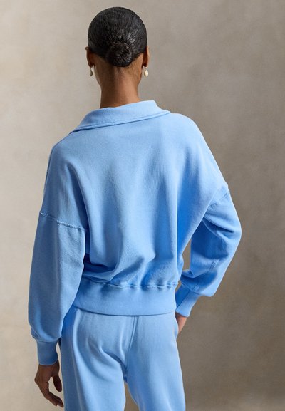 Γυναίκα με τα μαλλιά της σε κότσο φοράει ανοιχτό μπλε μπλούζα μακρυμάνικη και παντελόνι ταιριαστό, στέκεται με την πλάτη προς την κάμερα.