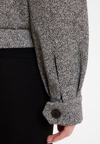 Wollen jas in een gemêleerd zwart-wit patroon, met een knoopsluiting bij de mouw en een geribbelde onderzoom. Close-up van het elleboogdetail.