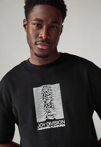 Black t-shirt featuring a white graphic depicting soundwave lines and the text "JOY DIVISION UNKNOWN PLEASURES" beneath. Fitted design.