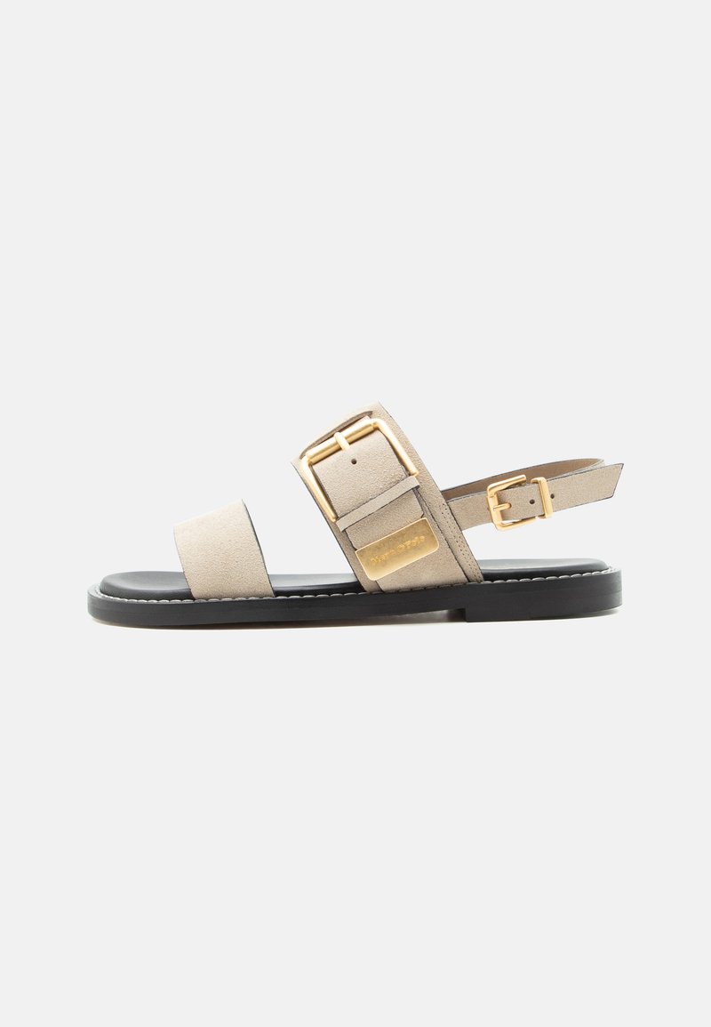 Béžová pásková sandála s černou podrážkou, širokým předním páskem, dvojitými zlatými přezkami a nastavitelným páskem kolem kotníku.
