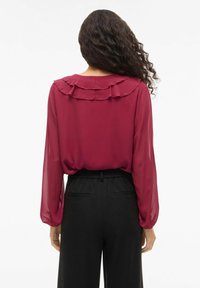 Bordowa bluzka z marszczonym kołnierzem, długimi prześwitującymi rękawami i luźnym krojem, w połączeniu z czarnymi, wysokim stanem spodniami z elastycznym pasem.