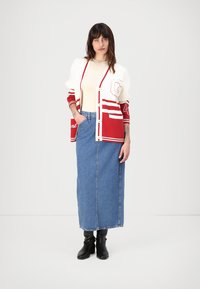 Rød og hvit varsity cardigan med knapper, preget av "G" og stripete detaljer, lagdelt over en kremfarget topp, kombinert med et denim maxiskjørt og svarte støvler.