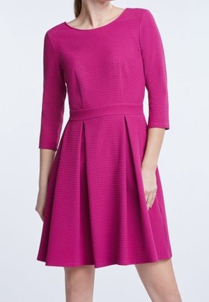 Kobieta w teksturowanej, jasnoróżowej sukience do kolan z rękawami do łokci i dopasowaną talią.