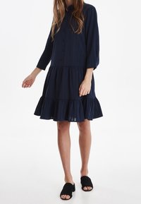 Abito blu navy con bottoni, orlato con volant a strati, maniche a tre quarti e una texture leggera, abbinato a mule nere con la punta aperta.
