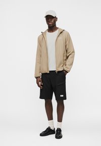 Άνδρας που φοράει μπεζ μπουφάν με κουκούλα, άσπρο καπέλο, άσπρο μπλουζάκι, μαύρες βερμούδες με τσέπες, άσπρες κάλτσες και μαύρα αθλητικά παπούτσια, στέκεται μπροστά σε λευκό φόντο.