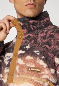 Close-up van een persoon die een bruine drukknopfleece kraag met een meerkleurig Columbia Sportswear Company logo en een rits op de borst aanpast.
