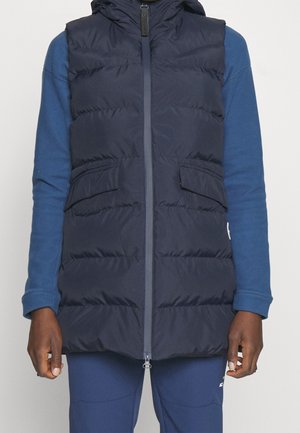 Personne portant une veste sans manches matelassée bleu marine par-dessus une chemise bleu à manches longues et un pantalon bleu assorti, debout devant un fond uni.