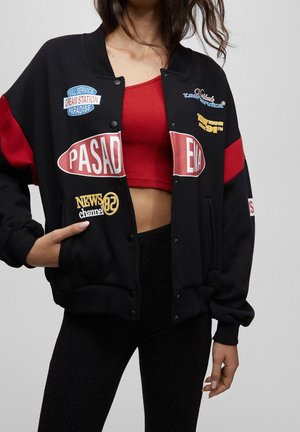Femme portant une veste bomber noire et rouge avec des patches sur un crop top rouge et un pantalon noir texturé, main dans la poche.