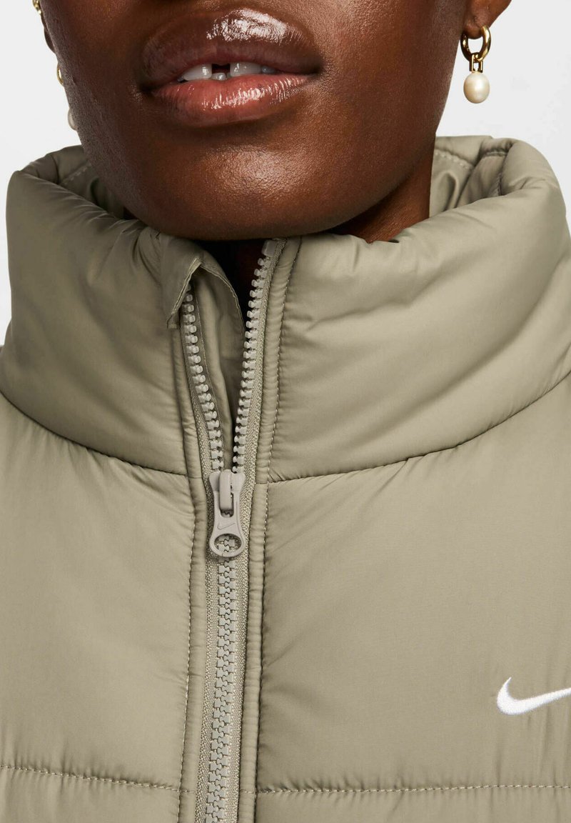 Giacca puffer in tessuto verde chiaro con colletto alto, chiusura con zip e logo Nike bianco; presenta una texture trapuntata.