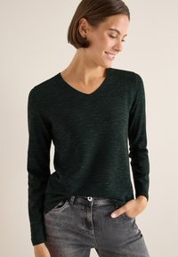 Dunkelgrüner Langarm-Pullover mit V-Ausschnitt, der ein strukturiertes Strickmuster aufweist. Kombiniert mit grauen Jeans für einen lässigen Look.