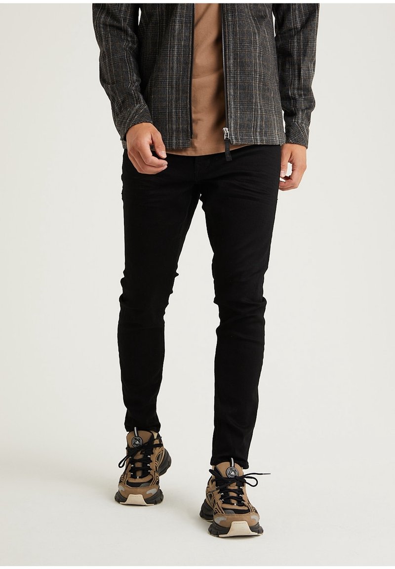 Pantalon noir fuselé en denim, porté avec une chemise marron et une veste sombre à motifs. Assorti avec des chaussures de sport marron et noires.