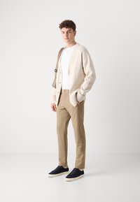 Cardigan bej tricotat cu manșete cu nervuri, tricou alb cu guler rotund, pantaloni bej și adidași bleumarin. Modelul stă în fața unui fundal simplu.