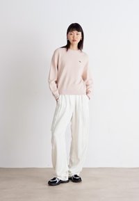 Suéter rosa claro com punhos e decote canelados, combinado com calças brancas largas. A modelo usa ténis pretos; design e corte minimalistas.