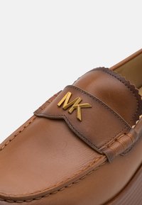 Pruunist nahast loafers, millel on kuldne MK logo, laineline detail ja tikanditega aktsent. Sile tekstuur ja ümar varvaste disain.