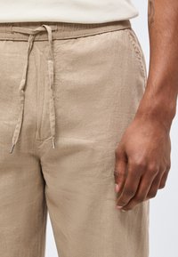 Κοντινό πλάνο ενός ατόμου που φοράει μπεζ λινά παντελόνια με κορδόνι στη μέση και το δεξί του χέρι ακουμπισμένο δίπλα στα παντελόνια.